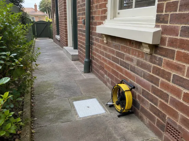Capped inspection opening on a paved side path beside a brick house and camera reel in Stanmore