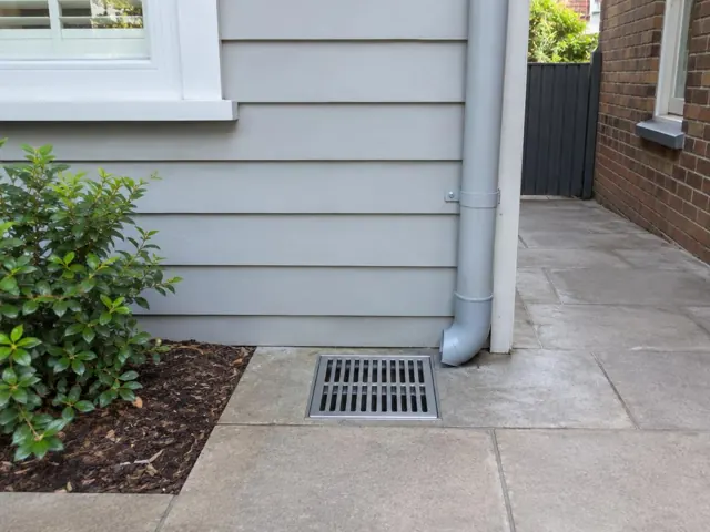 Outdoor metal drain grate beside grey weatherboard wall and brick path in Petersham