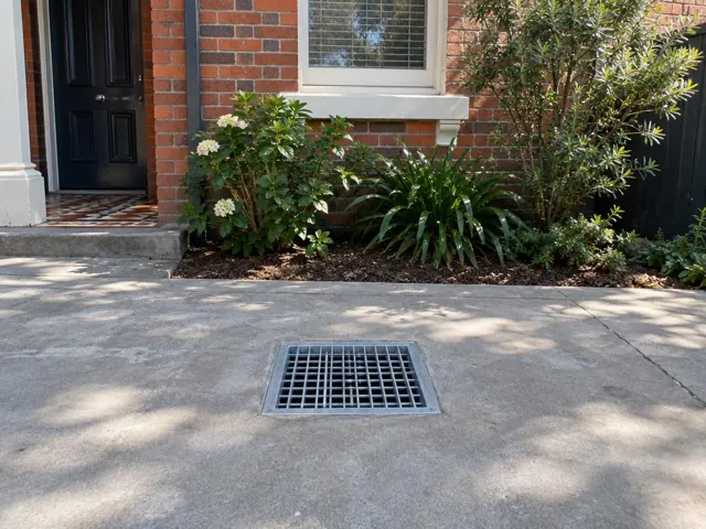 Concrete driveway with a seated stormwater grate beside a brick house and garden in Newtown